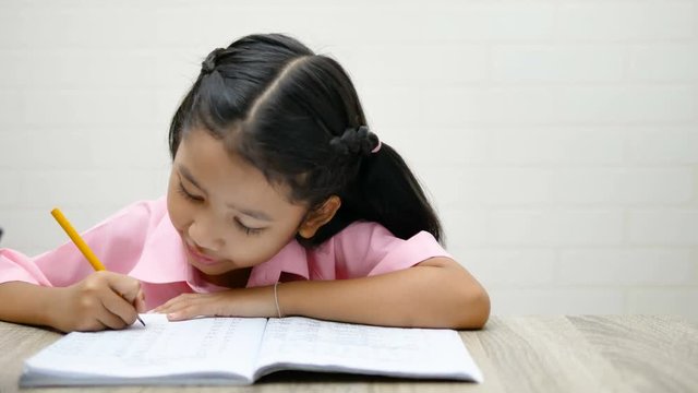 the little girl is doing homework happily. Children use a yellow pencil is writing a notebook on the wooden table.