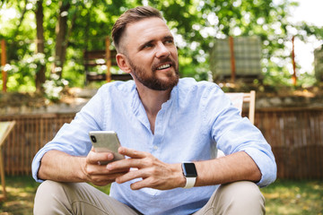 Naklejka na ściany i meble Happy young bearded man outdoors using mobile phone.