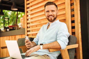 Smiling emotional young bearded man using laptop computer and mobile phone