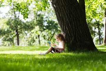 Little girl reading a book sitting on a grass under a tree summer day outdoors.
