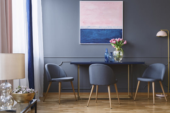Pink flowers on table in dining room interior with painting and grey chair next to lamp. Real photo
