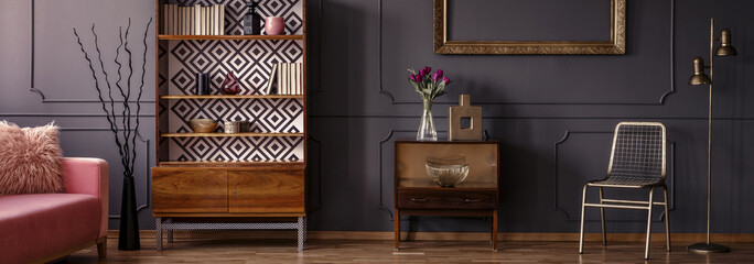 Gold chair standing in dark grey room interior with two vintage wooden cupboards with decor, books and fresh tulips