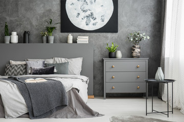 Monochromatic grey bedroom interior with a bed with linen and pillows. Decorative vases and plants above the bed, on a drawer cabinet and table by the window. Moon art on the wall. Real photo - obrazy, fototapety, plakaty