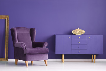 Purple cabinet with a golden vase, comfy armchair and frame in a living room interior