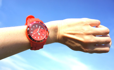 Female hand with red sports wrist watch on sky background. Five hours and five minutes. o'clock in...