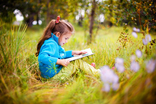 little girl in a blue sweater sits on the grass and reads a book