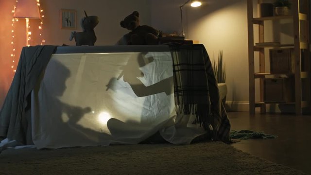 Silhouettes of two unrecognizable little children of elementary school age sitting behind piece of fabric and trying shadow play with flashlight
