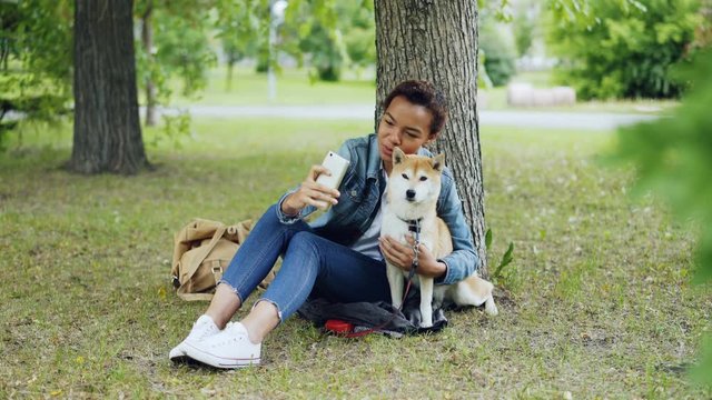 Pretty young girl blogger is taking selfie with purebred dog outdoors in city park cuddling and fondling beautiful animal. Modern technology, loving animals and nature concept.