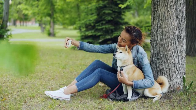 Attractive African American girl is taking selfie with cute dog resting in city park cuddling and caressing beautiful animal. Modern technology, loving pets and nature concept.