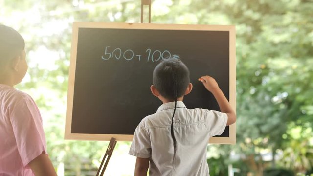 Asian boy two people who are creative, have fun learning math, writing answers to questions on the chalk board in black. Slow motion. tree nature background. Kid Creativity Education Concept....
