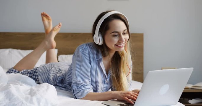 Young beautiful woman in the headphones lying on the unmade bed in the morning and chatting on the laptop computer, taping and typing. Inside