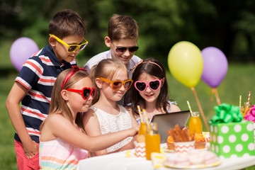 holidays, childhood and technology concept - happy kids in sunglasses with tablet pc on birthday party at summer garden