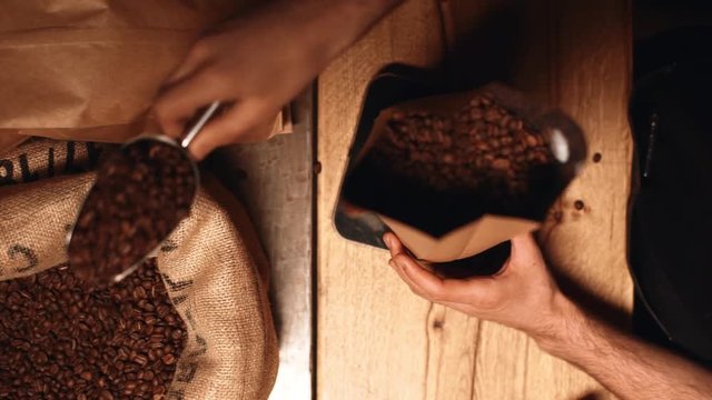 Closeup view from above in slow motion of male hand grabbing roasted robusta or arabica coffee beans from sack with metal shovel, and putting in paper bag for weighting on scale