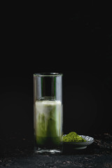 Matcha green tea iced latte or cocktail in tall glass with ice cubes and matcha powder over dark...