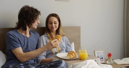 Portrait of the lovely young couple having breakfast with croissants and orange juice while sitting in the bed in the morning and man feeding his wife with a croissant. Indoor. - Powered by Adobe