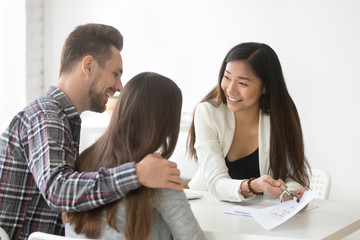 Smiling female real estate Asian talking about house plan to excited millennial couple, ready to buy first property together, spouses friendly meeting with broker, deciding to file for home mortgage