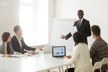 Obraz premium African American worker giving presentation on flipchart for diverse work group during business meeting, black coach mentoring mixed team talking about marketing plans and project success.