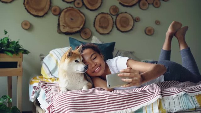 Happy African American girl proud dog owner is taking selfie with cute pet lying on bed in modern apartment using smartphone. Technology and social media concept.