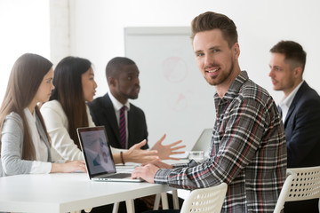 Portrait of smiling Caucasian male worker looking at camera while working at laptop at diverse team corporate business meeting, manager posing for photo during company briefing in office