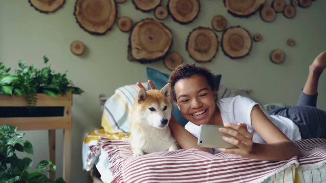 Cheerful girl is taking selfie with pet posing with beautiful dog lying on bed having fun and laughing. Modern apartment with lovely design and furniture is visible.