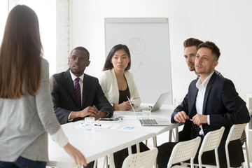 Diverse millennial team looking judgmental at female worker being late to meeting, angry colleague scolding unpunctual coworker for not coming on time to briefing. Punctuality, time management concept