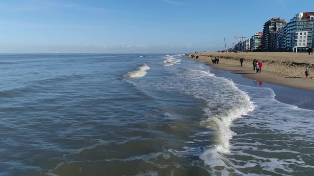 Low altitude aerial flight over surf showing the breaking waves and ocean on left and beach on right with people walking and playing on the relatively sunny but cold spring day 4k high resolution
