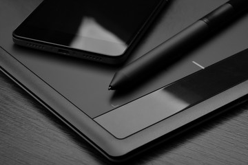 Smartphone and graphic tablet (also known as a digitizer or digital drawing tablet) with a special pen-like stylus on a wooden table. Details of workplace of an digital graphics designer