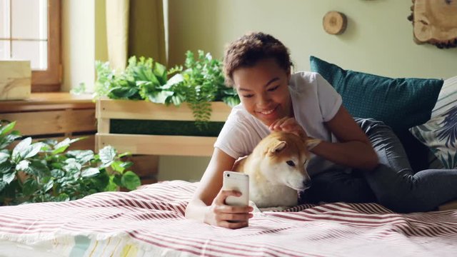 Joyful dog owner is taking selfie with cute pet lying on bed together holding smartphone posing and hugging animal. Friendship between people and puppies concept.