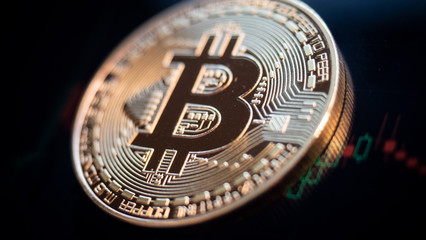Close up of a golden glowing bitcoin. In the background, are candle stick graphs and courses on crypto stock exchanges. BTC coin is the symbol of decentralized cryptocurrency based on blockchain.