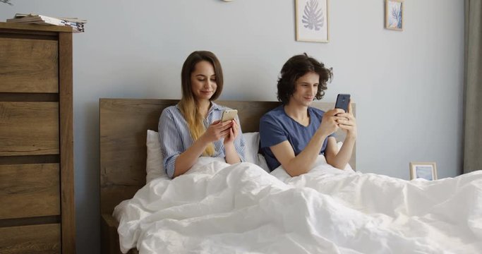Caucasian attractive couple sitting in the bedroom in the morning, taping on their smartphones and playing. Inside