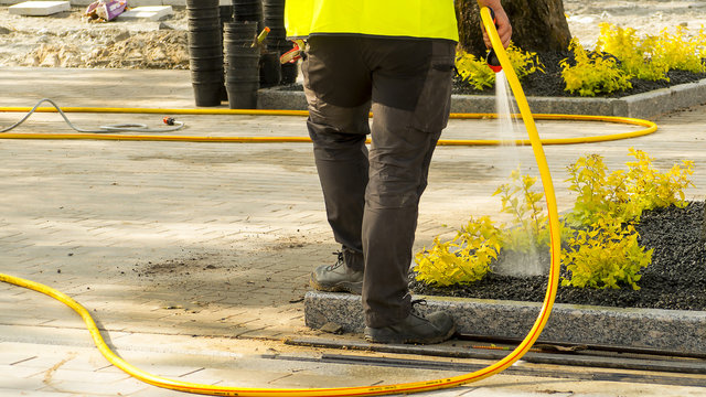 a street worker female in work clothes watering a new road and a lawn in the park with a hose-pipe. Tallinn, Estonia