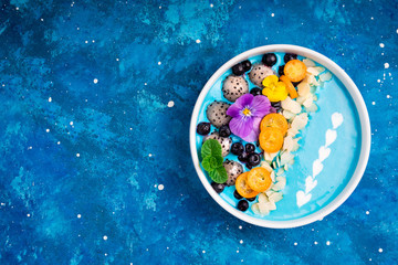 Blue smoothie bowl with fruits, berries, nuts and flowers. Tropical healthy smoothie dessert. Healthy food, vegetarian, diet concept. Top view. Copy space