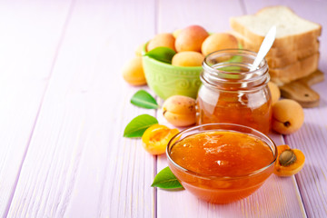Apricot jam in glass bowl and fresh apricots on light purple wooden background