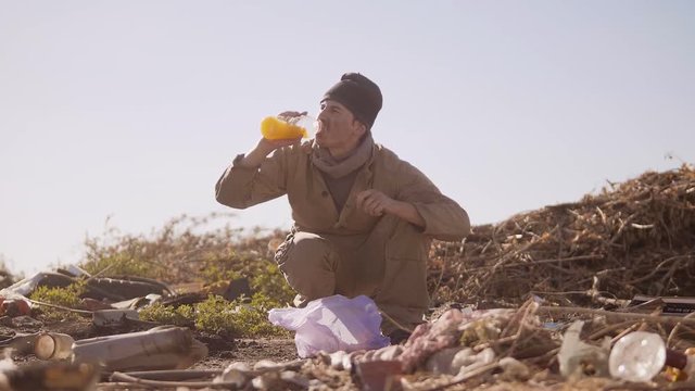 dirty homeless hungry man in a dump drinks the missing juice in the package with walking goes looking for food slow motion video. homeless dirty man roofless person looking for food in a dump