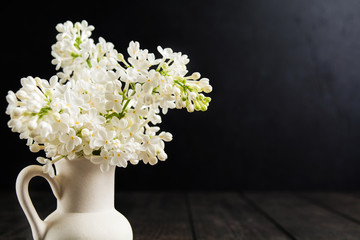 White lilac in ceramic jar over wooden table with black wall background with copy space