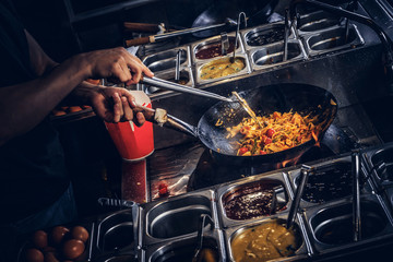 Cooking process in an Asian restaurant. Cook is stirring vegetables in wok. - obrazy, fototapety, plakaty