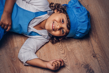 Cute little boy with brown curly hair dressed in a blue cook uniform lying on a wooden floor with fruits.