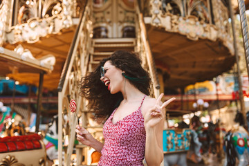 Cheerful woman with dark curly hair in sunglasses and dress standing with lolly pop candy in hand and happily looking aside while spending time in amusement park