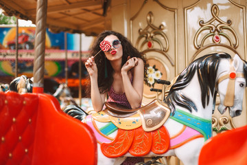 Young pretty lady with dark curly hair in sunglasses covering her eye with lolly pop candy while leaning on carousel hourse in amusement park