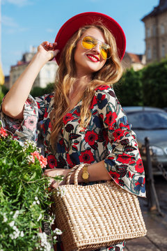 Outdoor portrait of young beautiful happy smiling woman wearing stylish jumpsuit with floral print, red hat, yellow sunglasses, wrist watch, holding straw bag. Model posing in street of european city