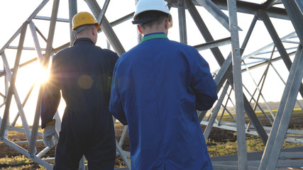 Fototapeta premium An electrician or builder and engineer, in a blue robe, in a yellow and white helmet, check the technical overview for strength, prepare electric poles, an electric tower, energy, and workers.