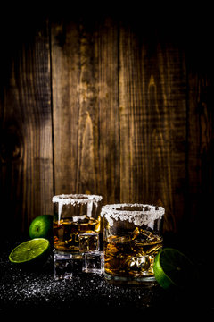 Two tequila shot glasses on dark background, with ice cubes, salt and limes copy space