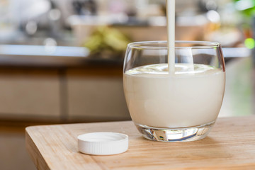 Hand pouring fresh kefir probiotik drink from white bottle into clear glass on kitchen table