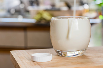 Hand pouring fresh kefir probiotik drink from white bottle into clear glass on kitchen table