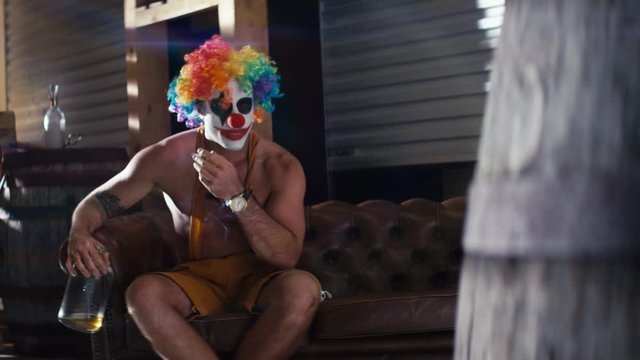 Clown in overalls and colorful wig smoking on couch and drinking