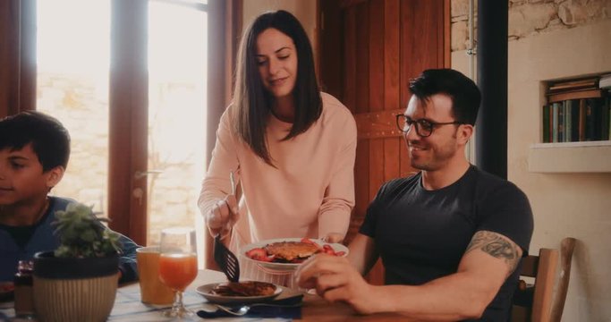 Wife serving husband pancakes during family breakfast in house kitchen