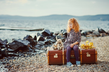 A little sad girl with a toy sitting on a large suitcase near th