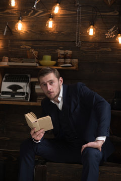 Enjoy moment. Man with beard holds old book and read. Guy in cozy warm atmosphere relaxing while reading. Man in formal suit enjoy evening with favourite literature, wooden background