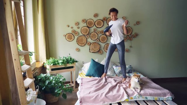 Playful young lady is singing in blow dryer, dancing and jumping on bed and listening to music while pet dog is lying on bed in nice modern style apartment.