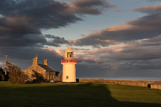 Ballinacourty lighthouse lit by the setting sun with the sea and dark clouds in the background. Ballinacourty, County Waterford, Republic of Ireland.